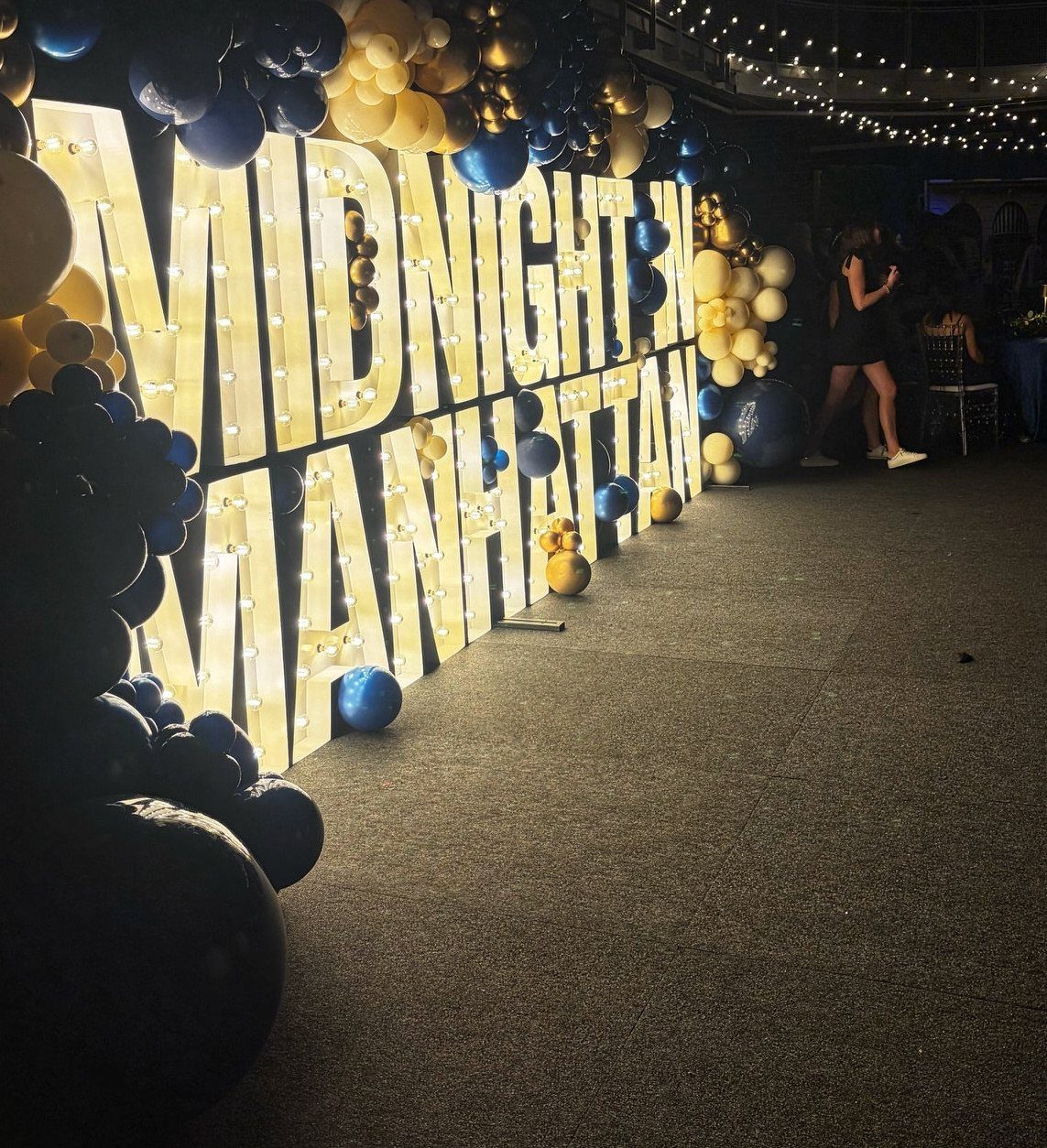 Illuminated Midnight in Manhattan marquee letters surrounded by gold and navy blue balloons.