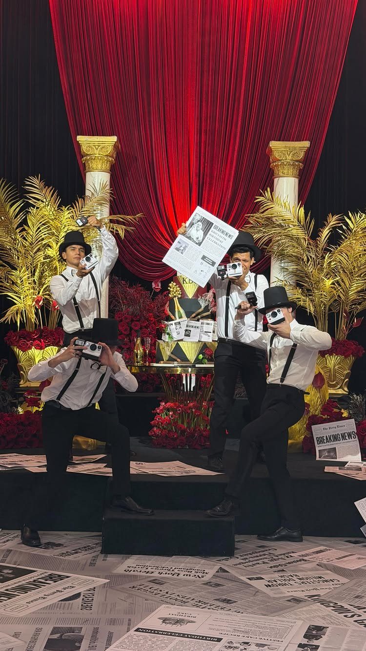 Paparazzi in vintage attire posing before red curtains and gold columns at an event.