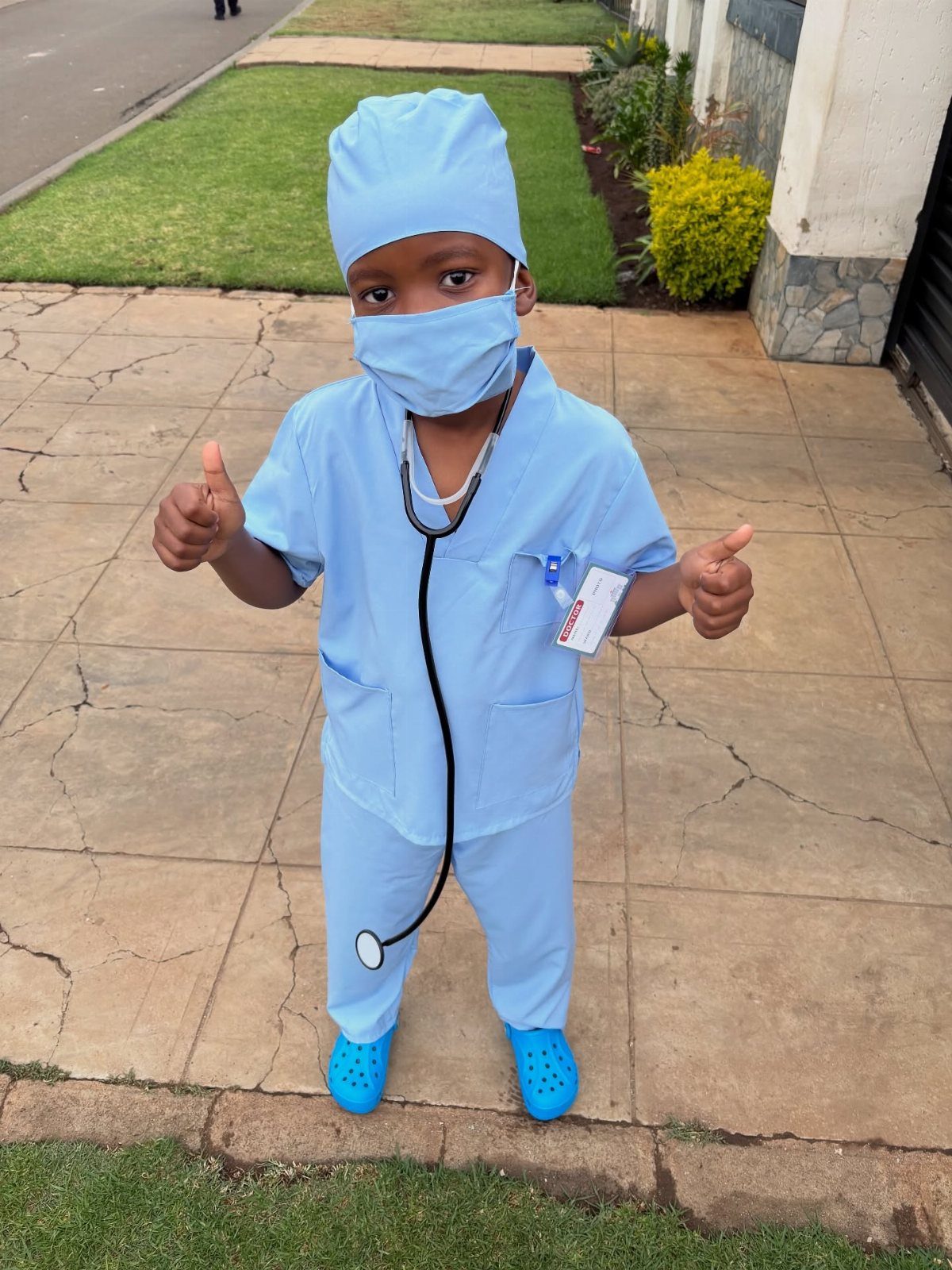 Young child dressed as a doctor in blue scrubs and stethoscope giving thumbs up.