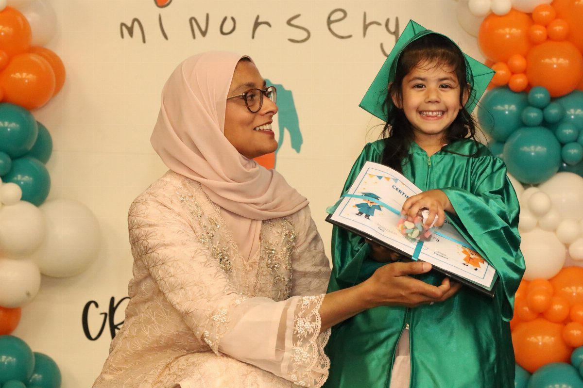 Young girl in green graduation gown holding a certificate with a smiling woman.