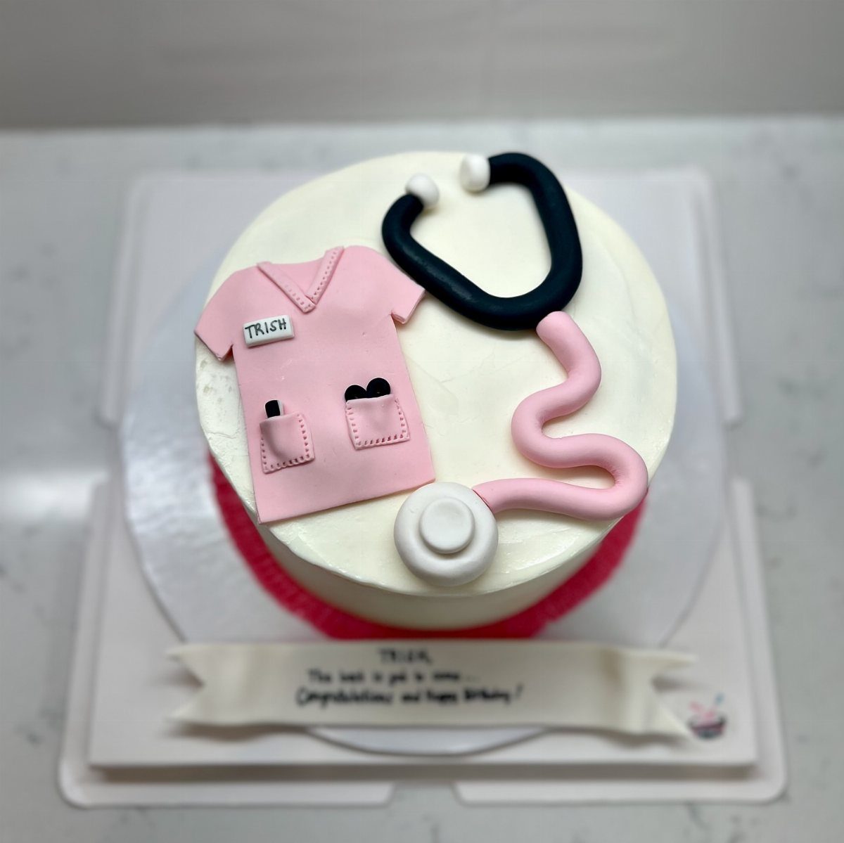 White frosted cake decorated with pink nurse scrubs and a black stethoscope.