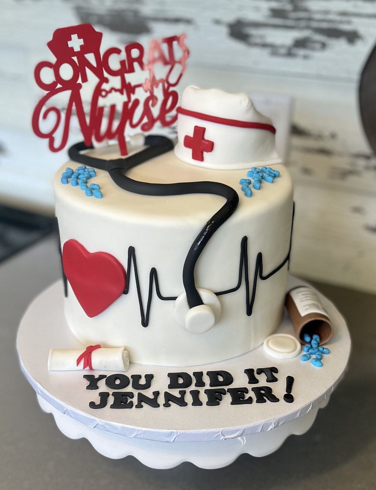 White nursing graduation cake topped with a nurse's hat, stethoscope, and diploma scroll.