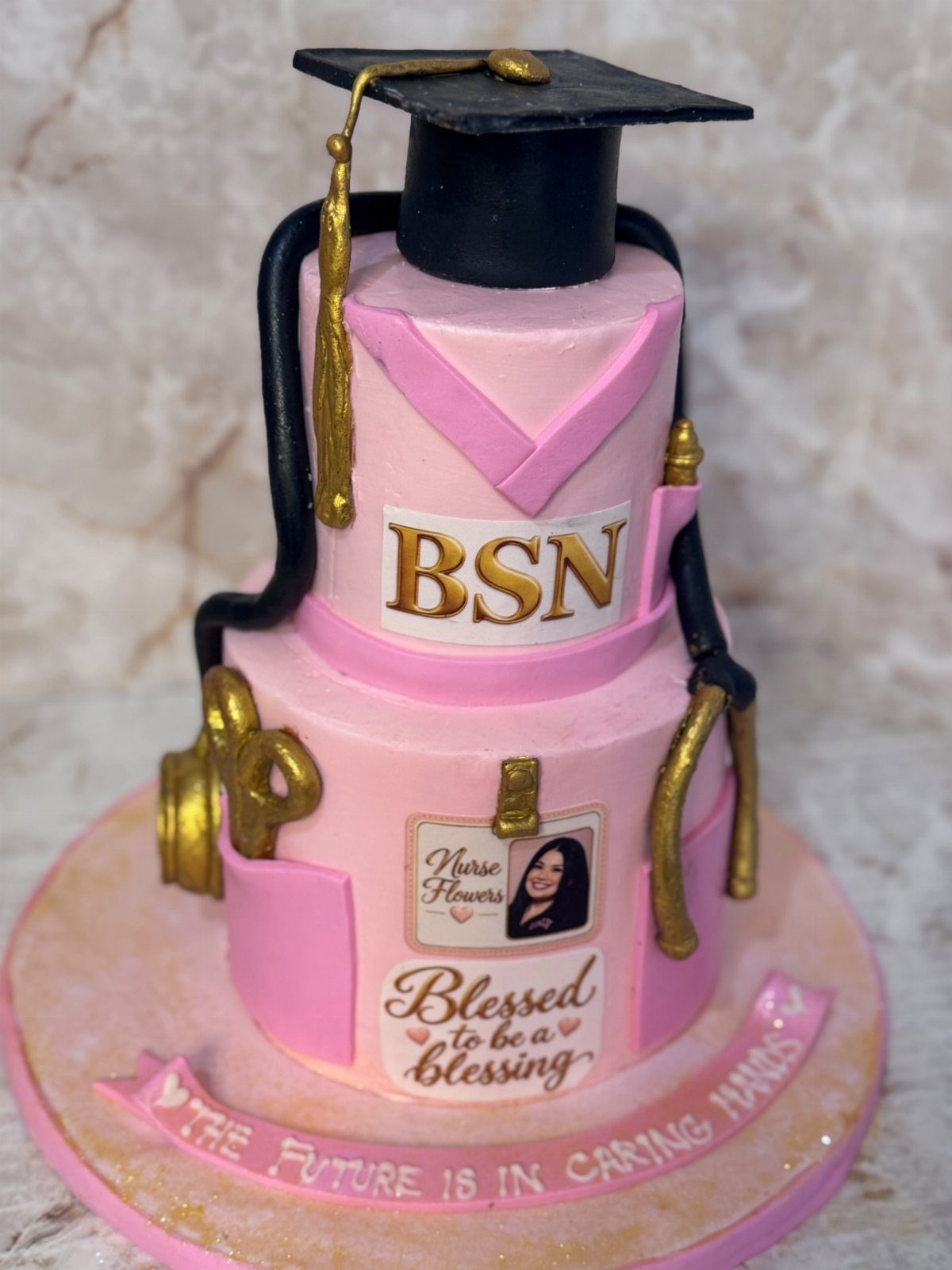 Two-tier pink nursing graduation cake with a black cap and gold stethoscope.
