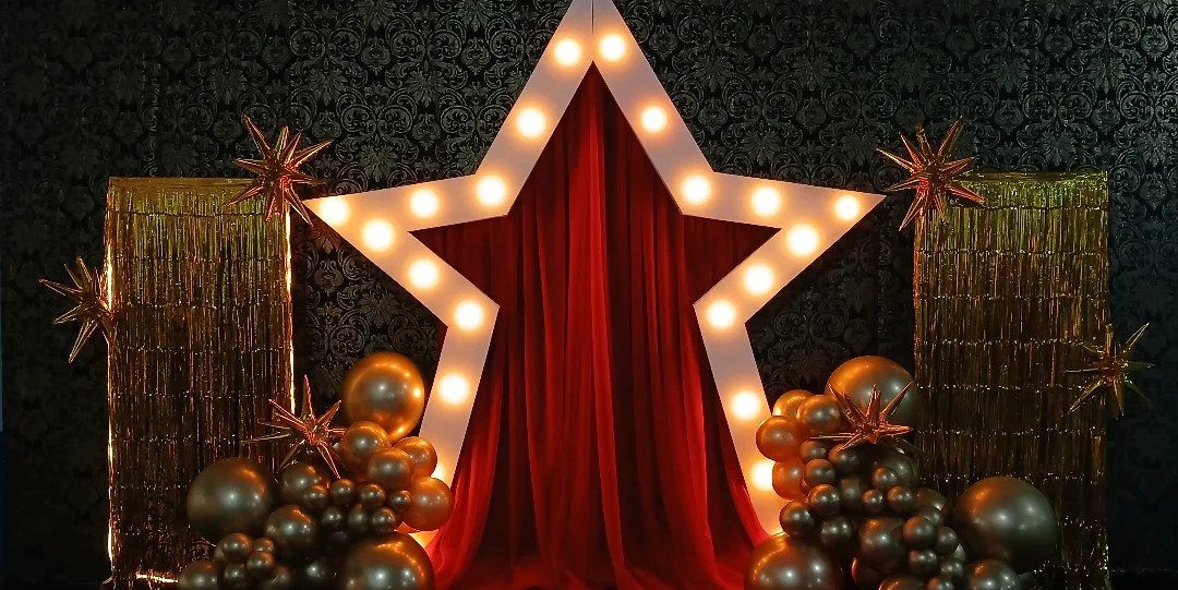 Large illuminated star frame with red curtains, gold balloons, and tinsel backdrop.