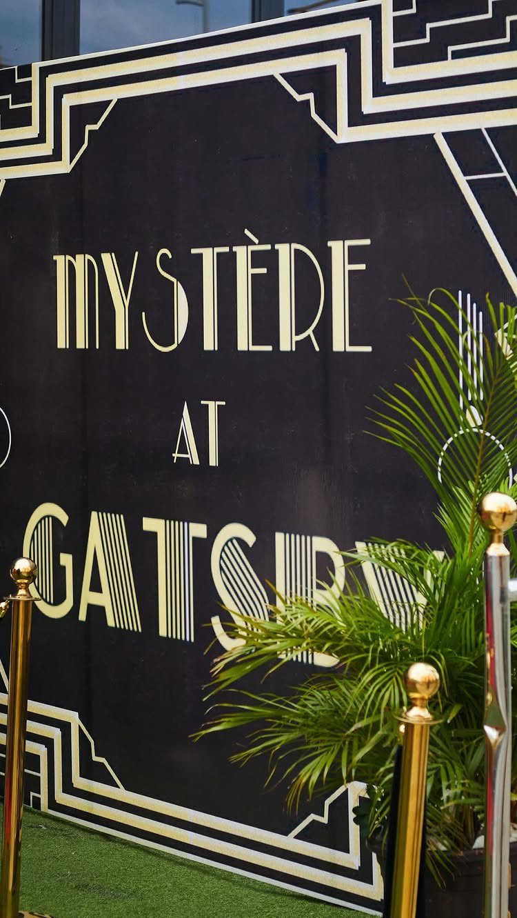 Black and gold Art Deco backdrop with Gatsby text, gold stanchions, and palm leaves.