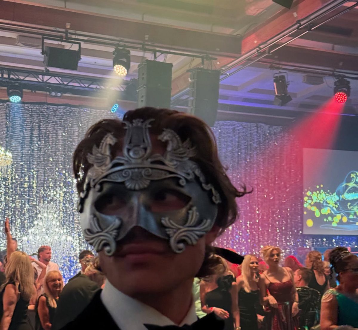 Young man in a tuxedo wearing an ornate silver masquerade mask at a party.