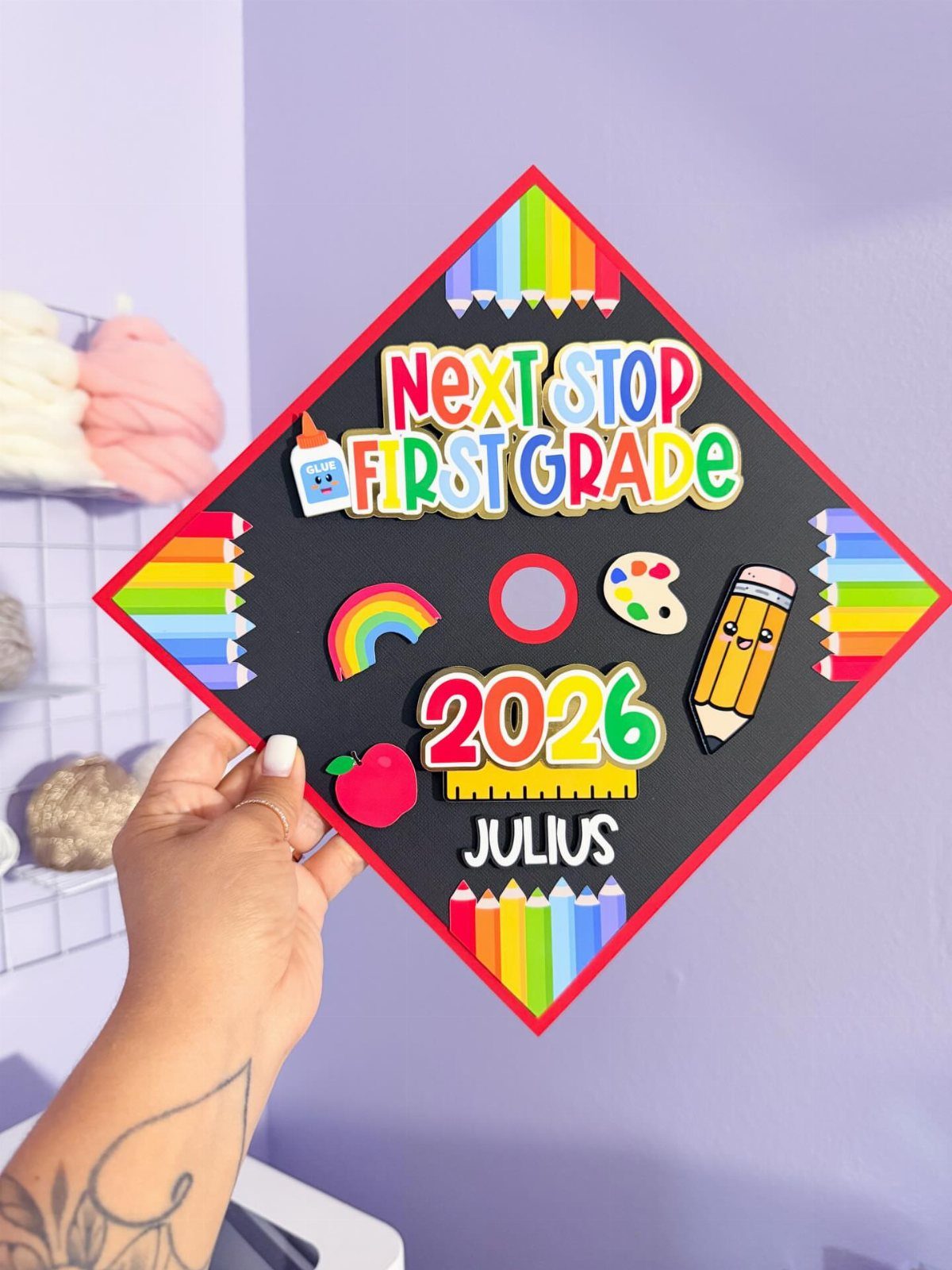 Decorated black graduation cap with colorful school-themed cutouts and text for first grade.