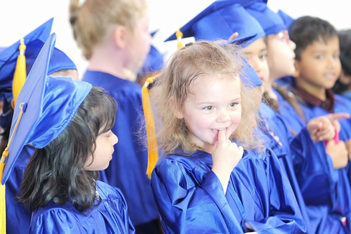 Young children in blue graduation gowns and caps smiling during a ceremony.
