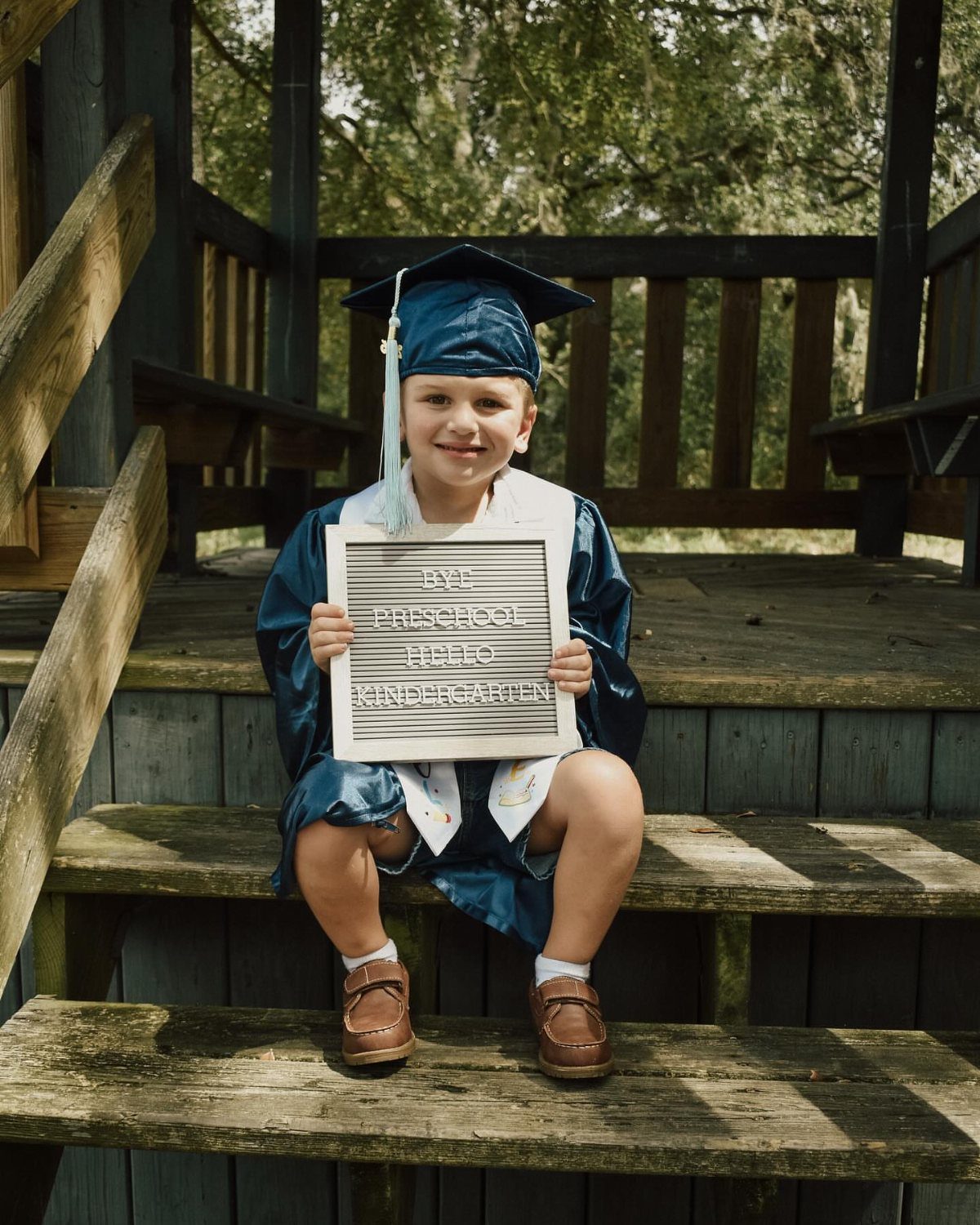 Young boy in blue graduation gown holding a preschool graduation sign on stairs