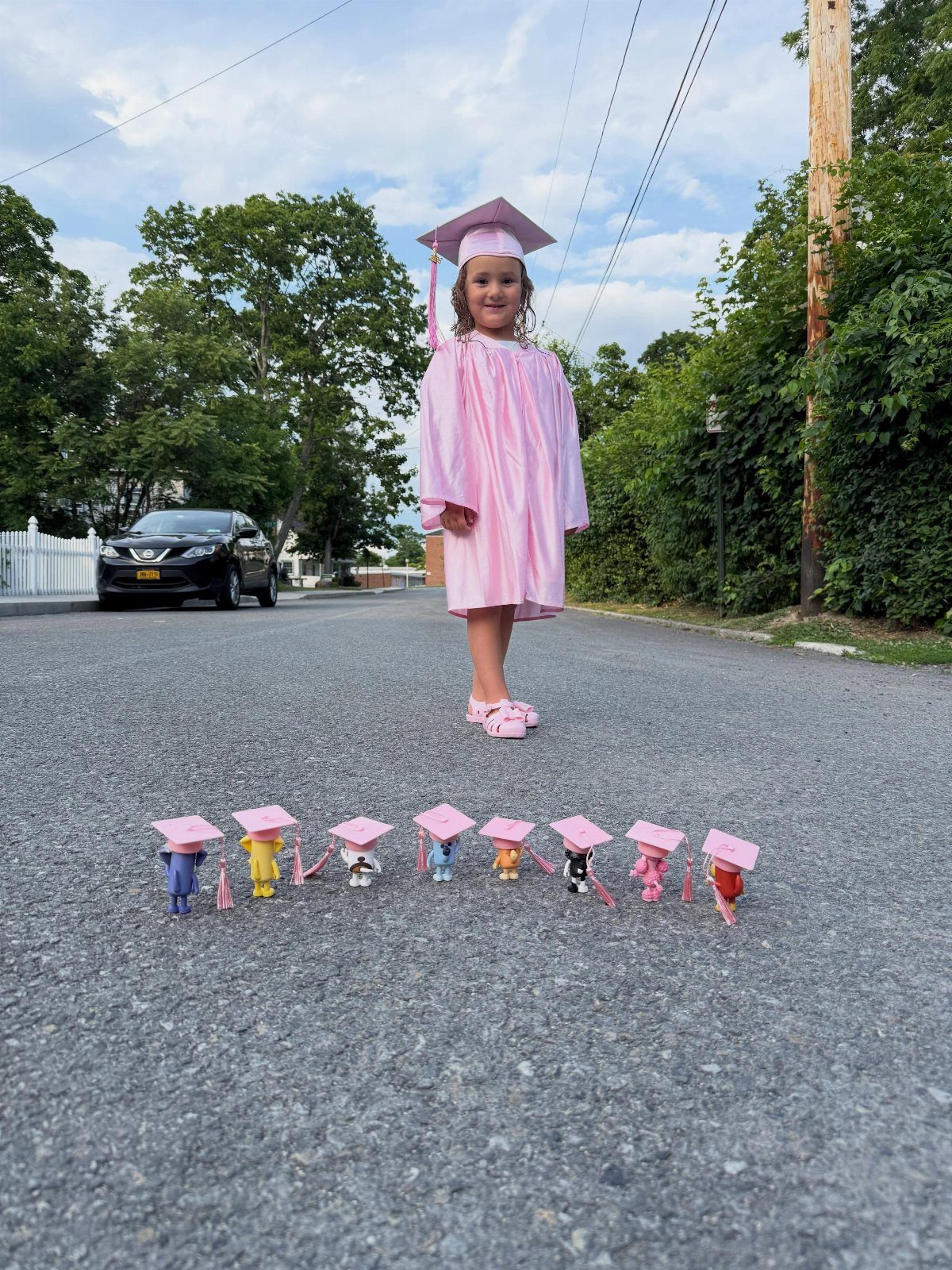 Young girl in pink graduation cap and gown standing with toy graduates.