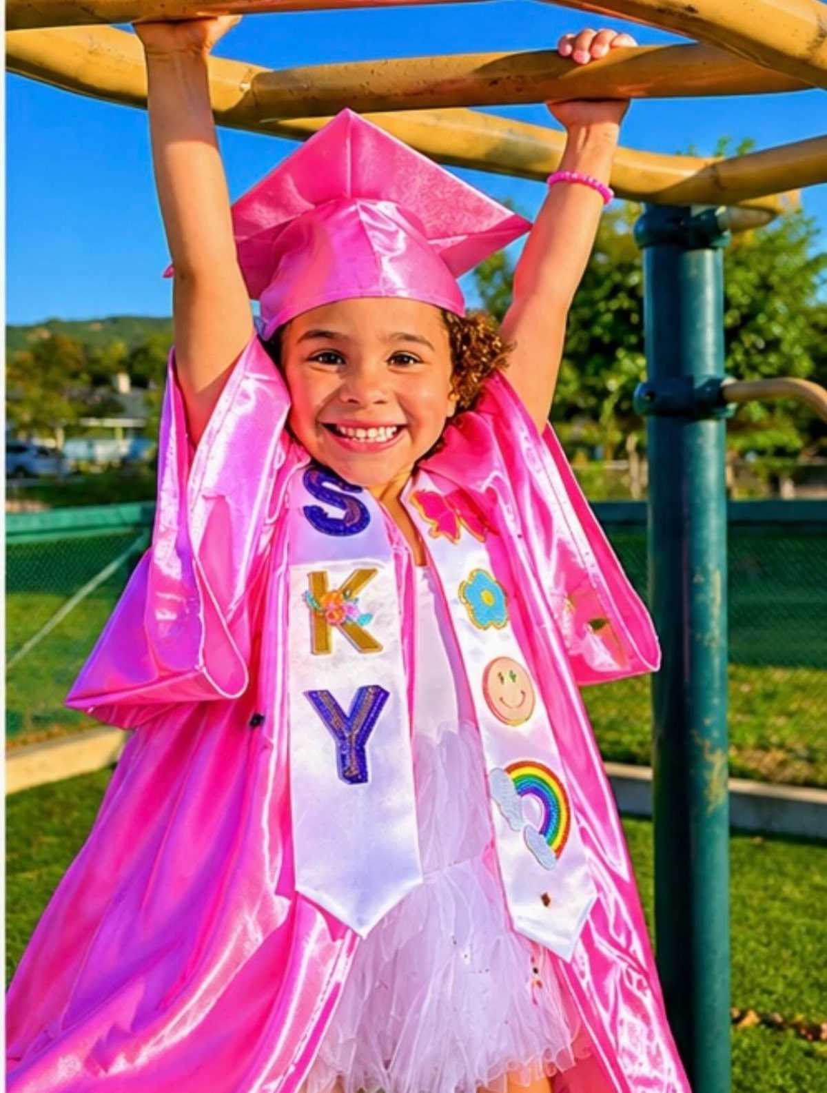 Young girl in pink graduation cap and gown smiling on playground monkey bars