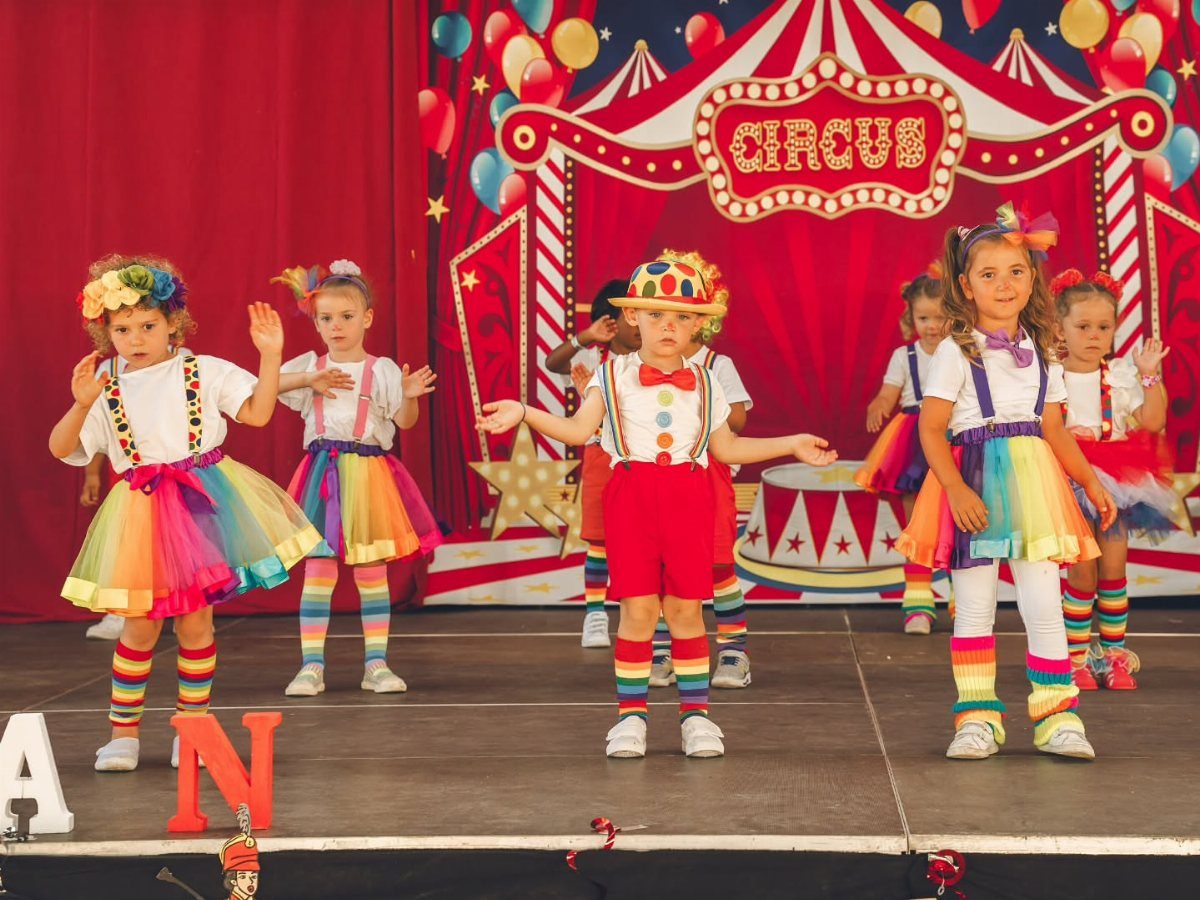 Children in colorful circus costumes posing on a stage with a circus backdrop.