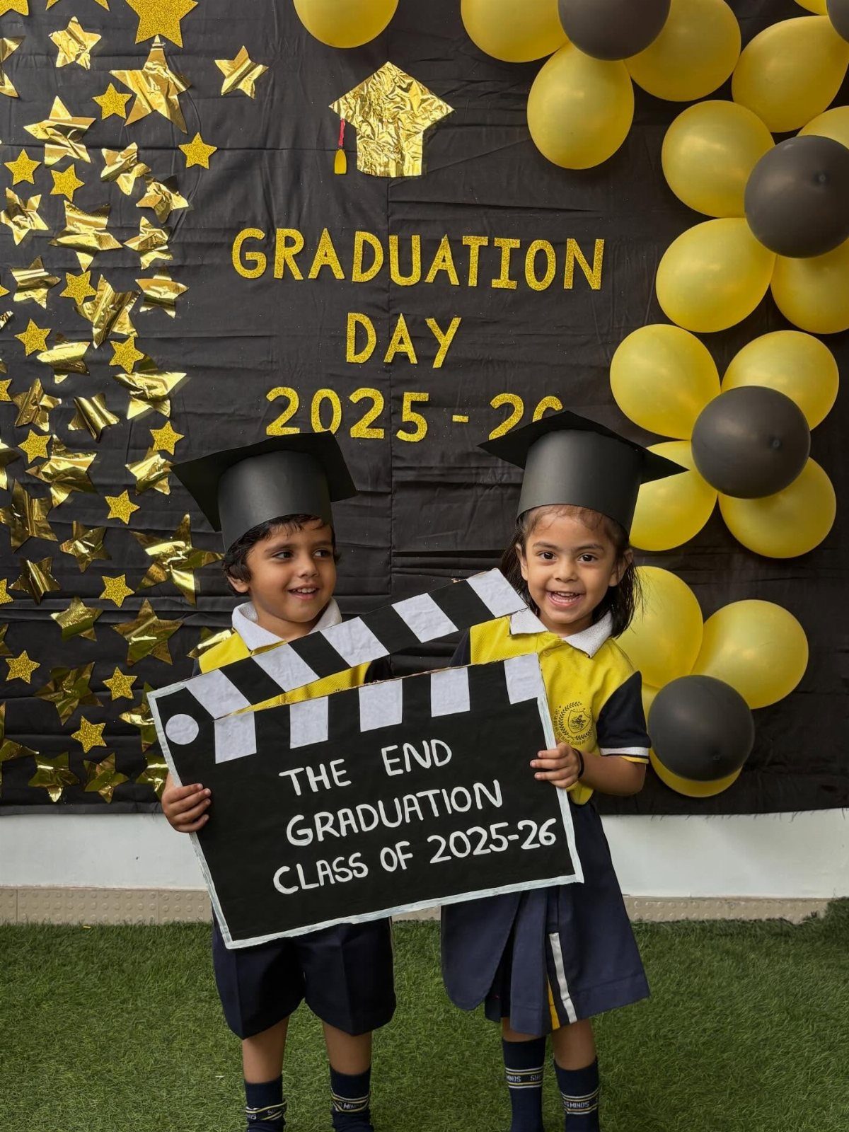 Two preschool graduates in caps holding a movie clapperboard against a balloon backdrop.