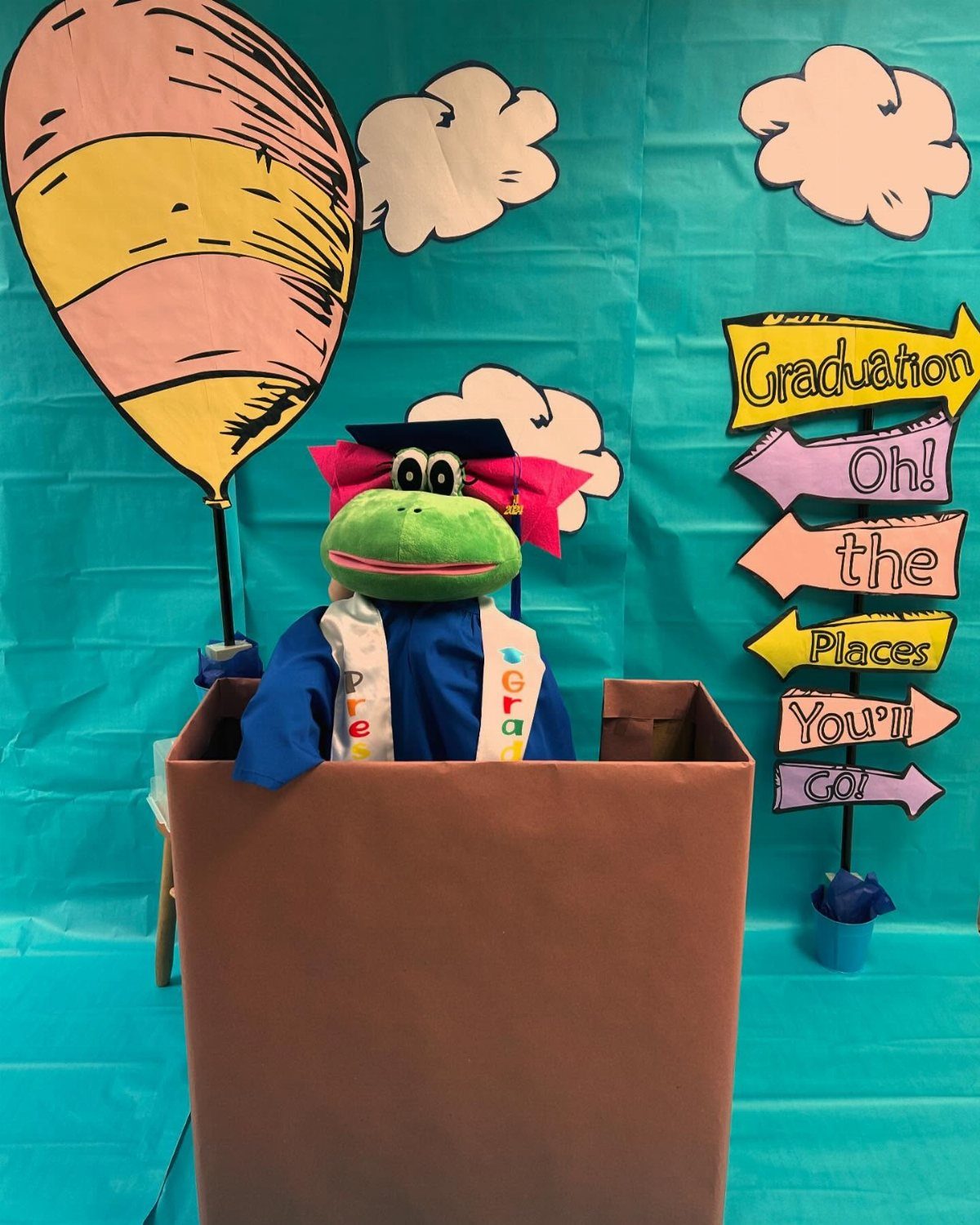 Graduation frog plush in a box with Dr. Seuss themed backdrop and signs.