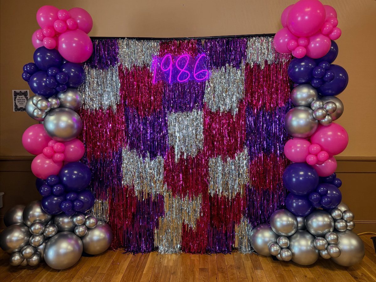 Party backdrop with pink, purple, and silver balloons and a neon 1986 sign.