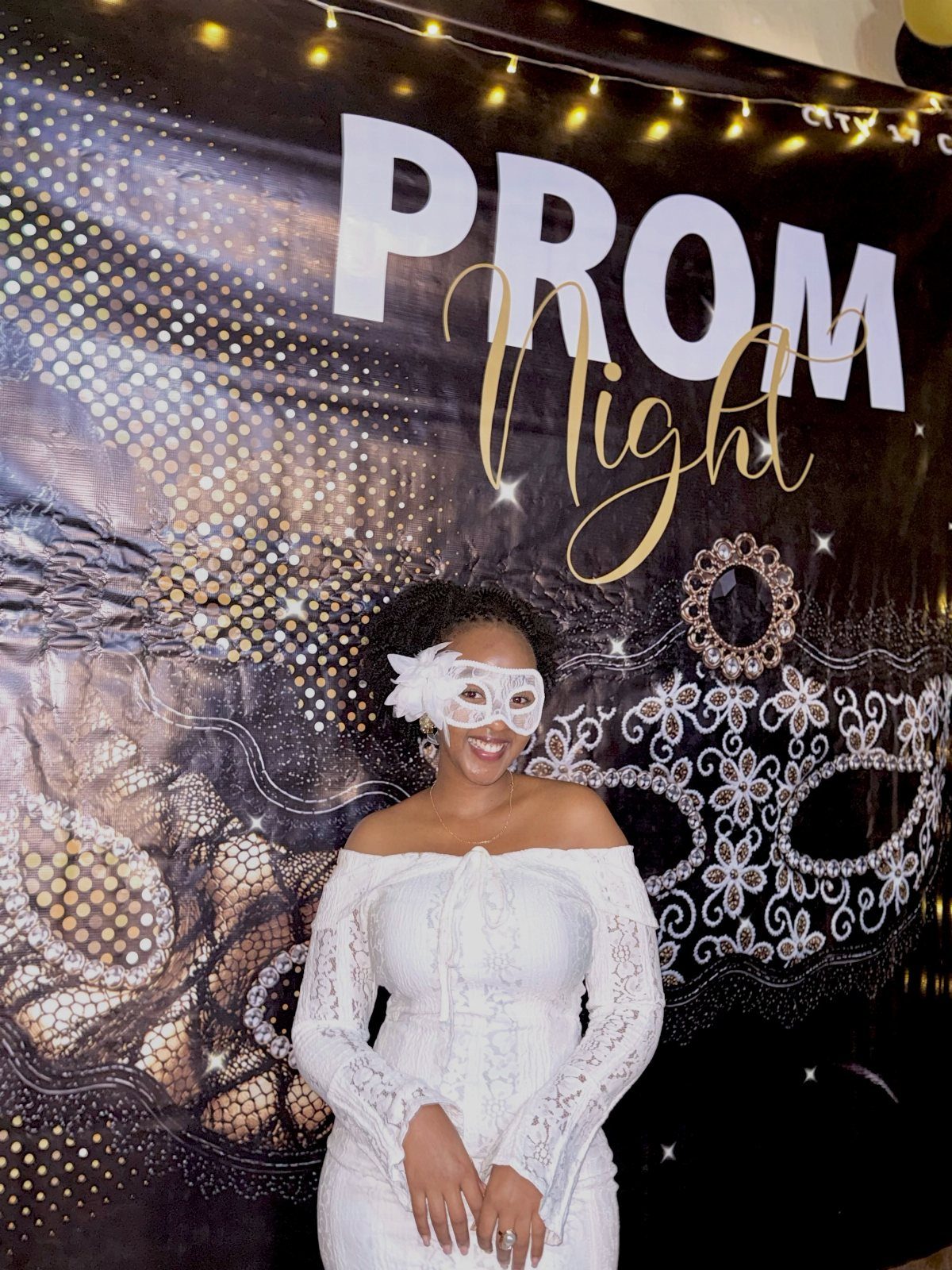 Woman in white lace dress and masquerade mask posing before prom backdrop