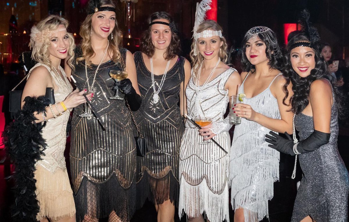 Six women in 1920s flapper dresses and headbands posing with cocktails at a party.