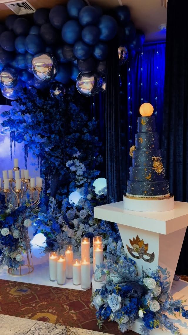 Blue cake with gold accents surrounded by blue flowers, balloons, and white candles.
