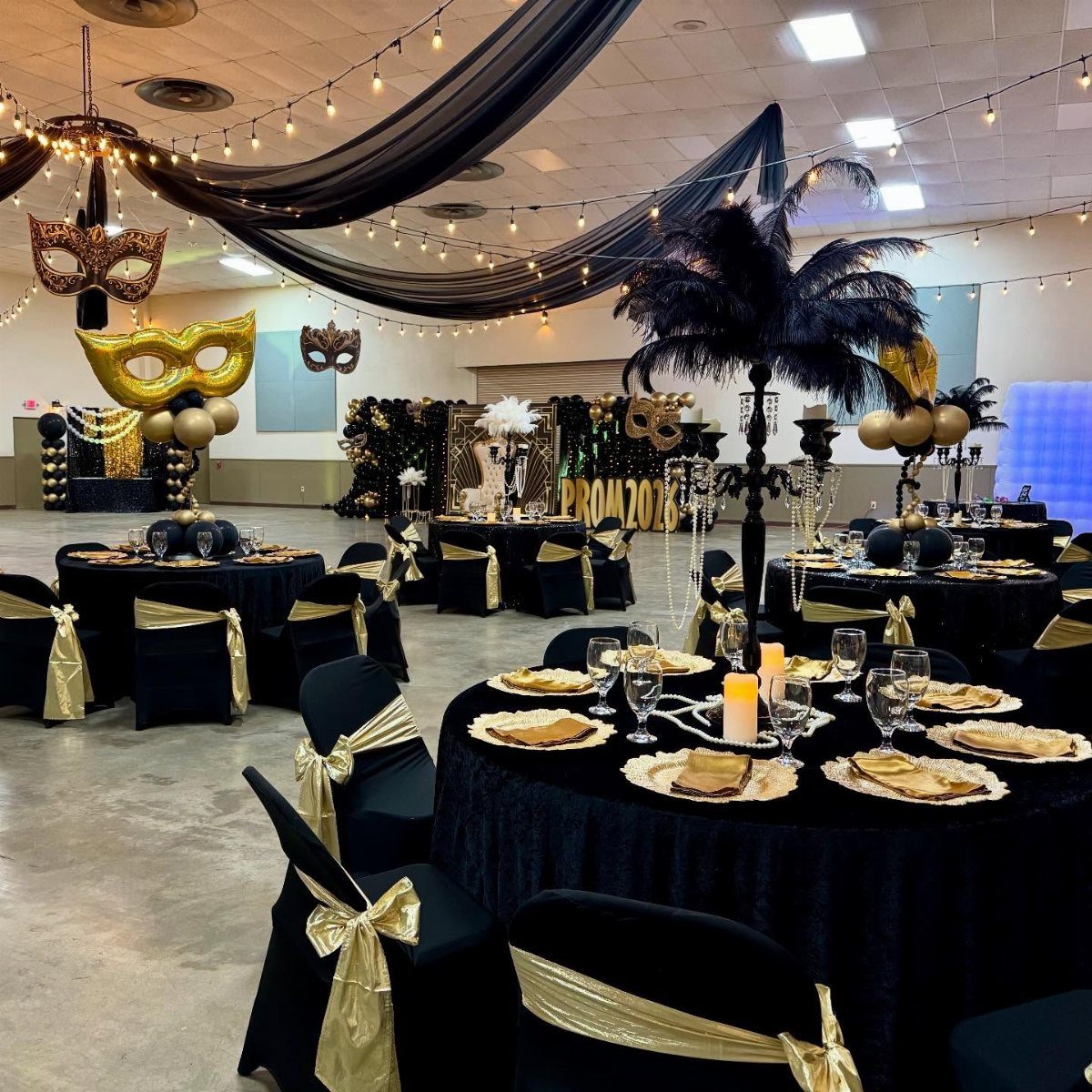 Banquet hall with black and gold decor featuring large masquerade masks and string lights.