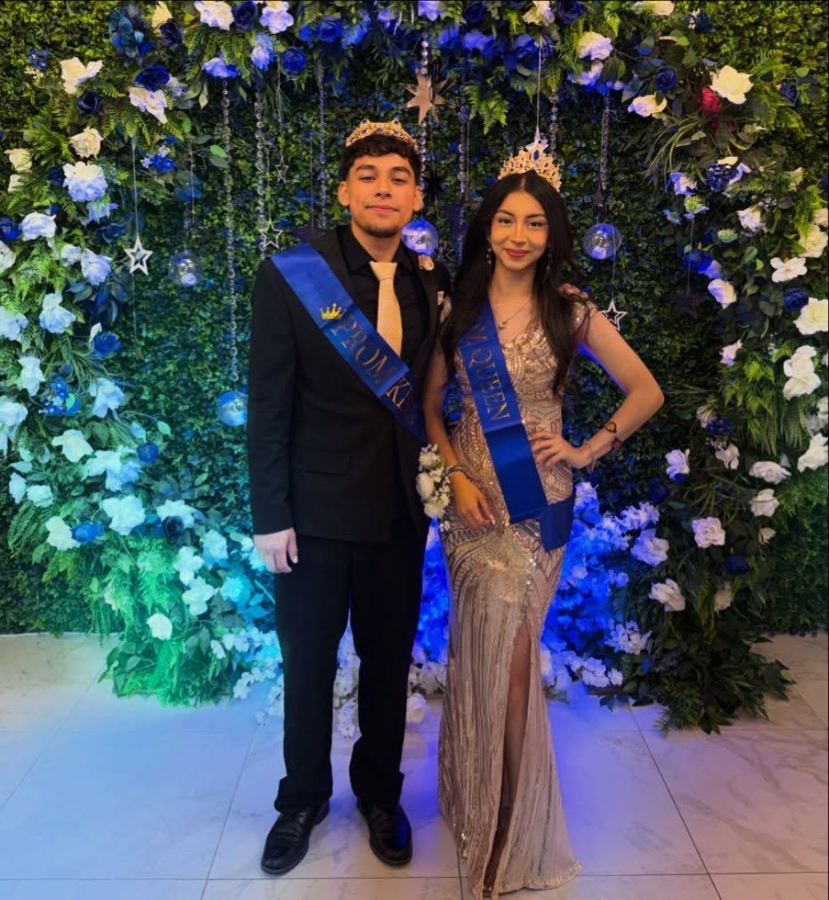 Prom king and queen wearing crowns and blue sashes posing before floral wall.