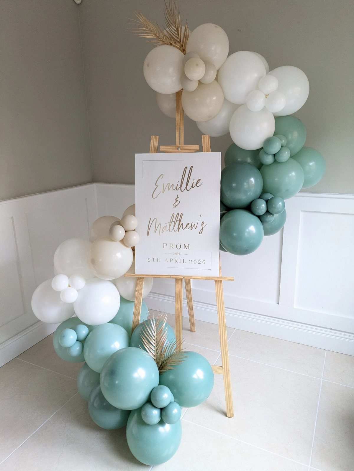 Custom prom welcome sign on a wooden easel surrounded by sage and white balloons.