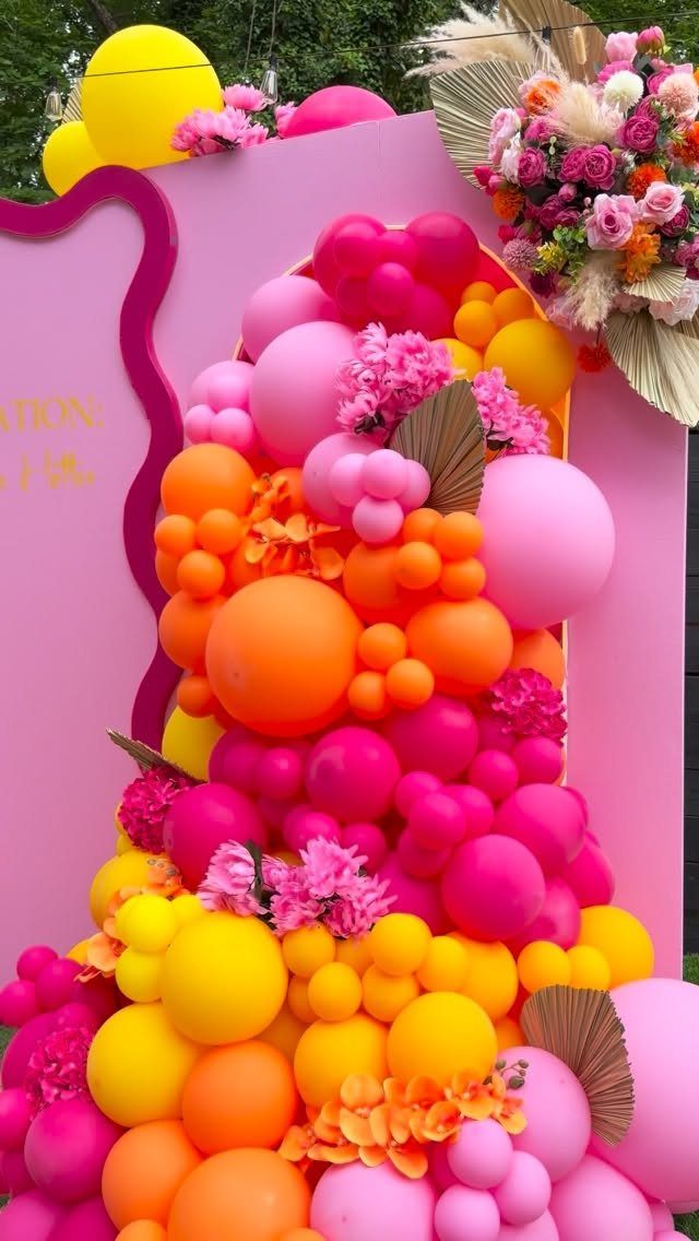 Colorful balloon arch with bright pink and orange flowers and dried palm leaves.