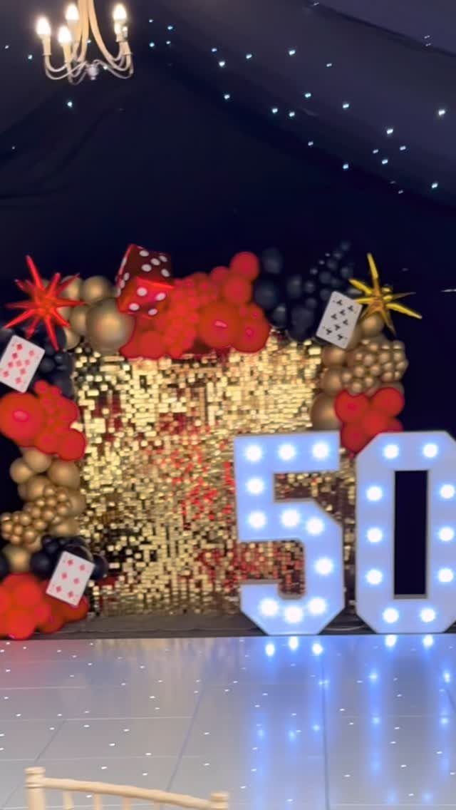 Gold sequin backdrop with red and black balloons and large glowing number 50