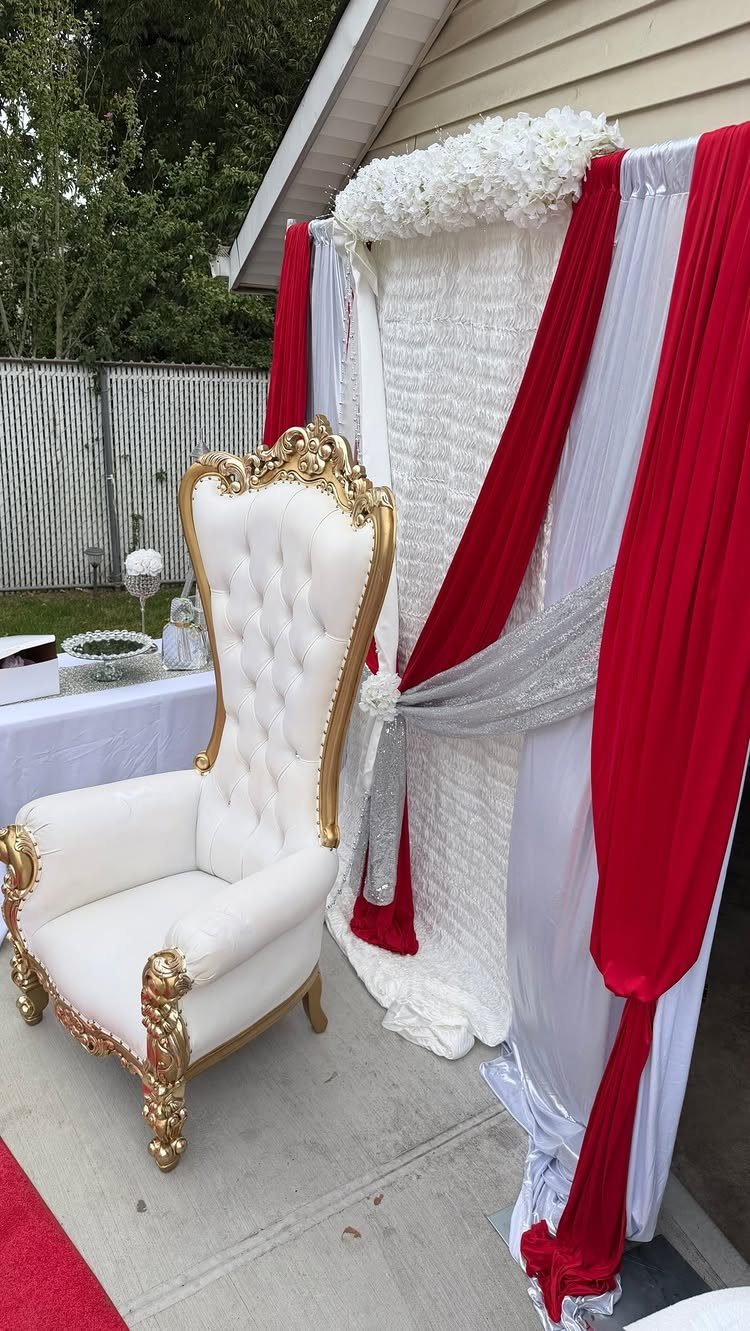 Ornate white and gold throne chair beside red and white draped fabric backdrop.