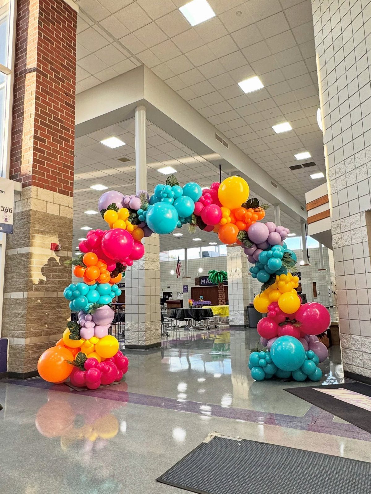 Colorful balloon arch with tropical leaves in a bright school hallway.