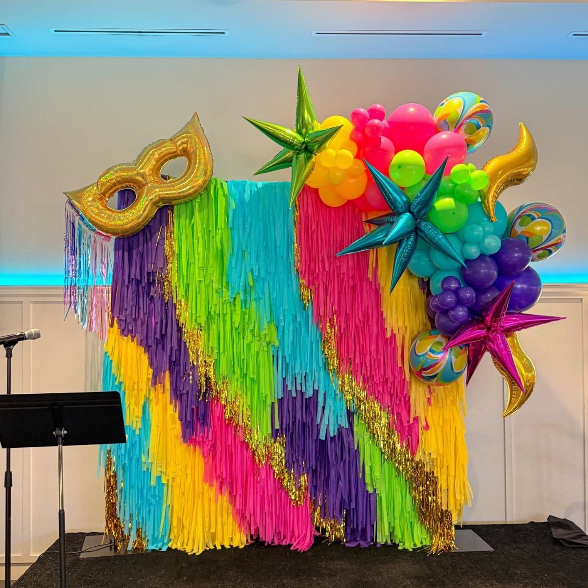 Colorful streamers backdrop with gold mask and vibrant balloon decorations for a party.