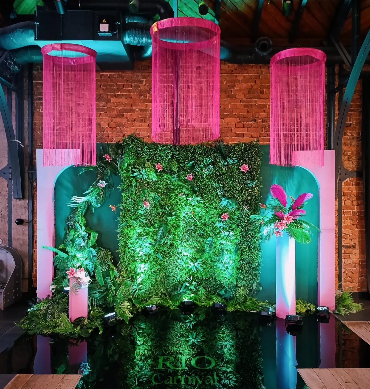 Lush green foliage wall with pink tropical flowers and hanging pink fringe decorations.