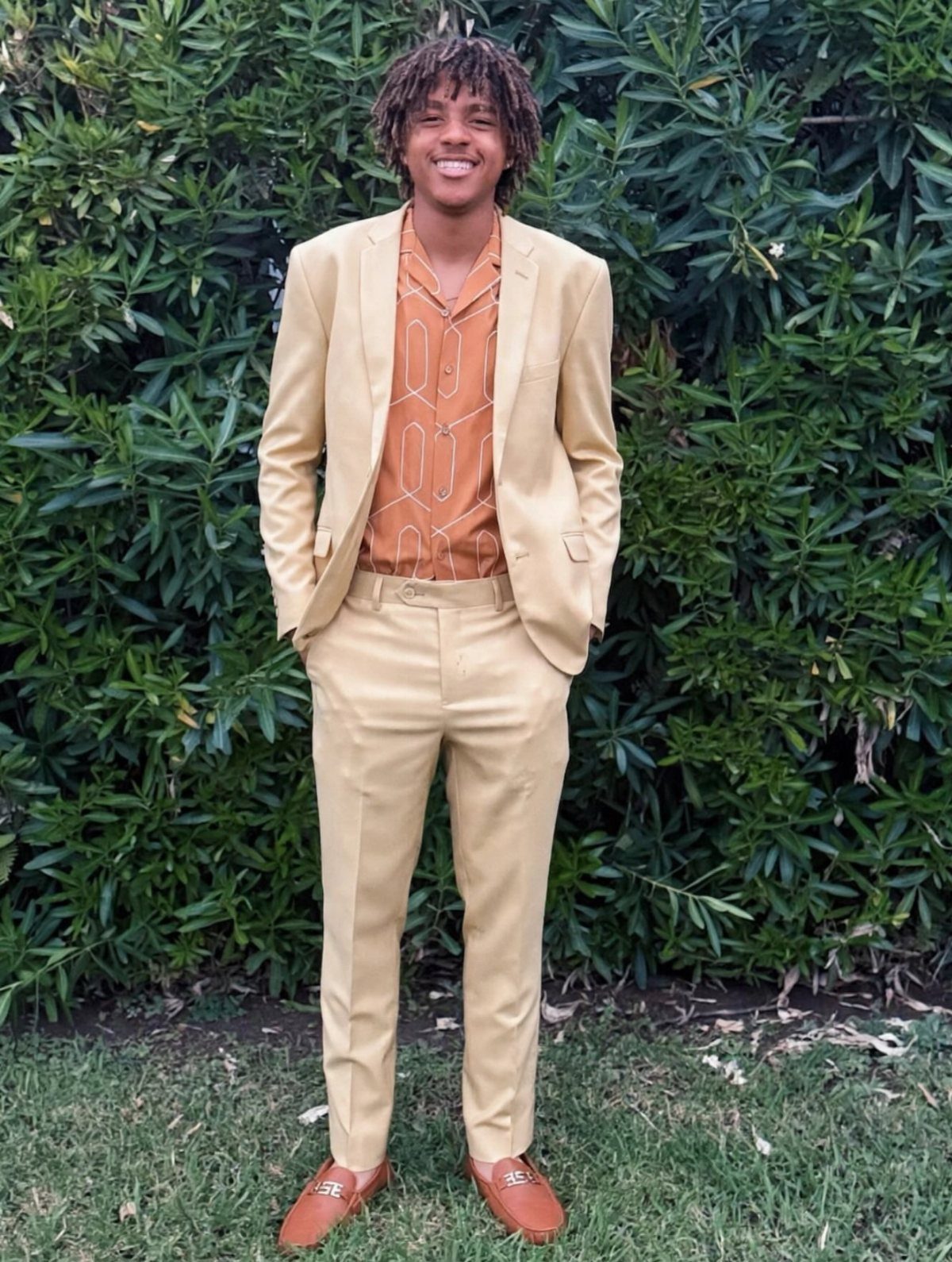 Young man wearing a tan suit and orange patterned shirt against green bushes.