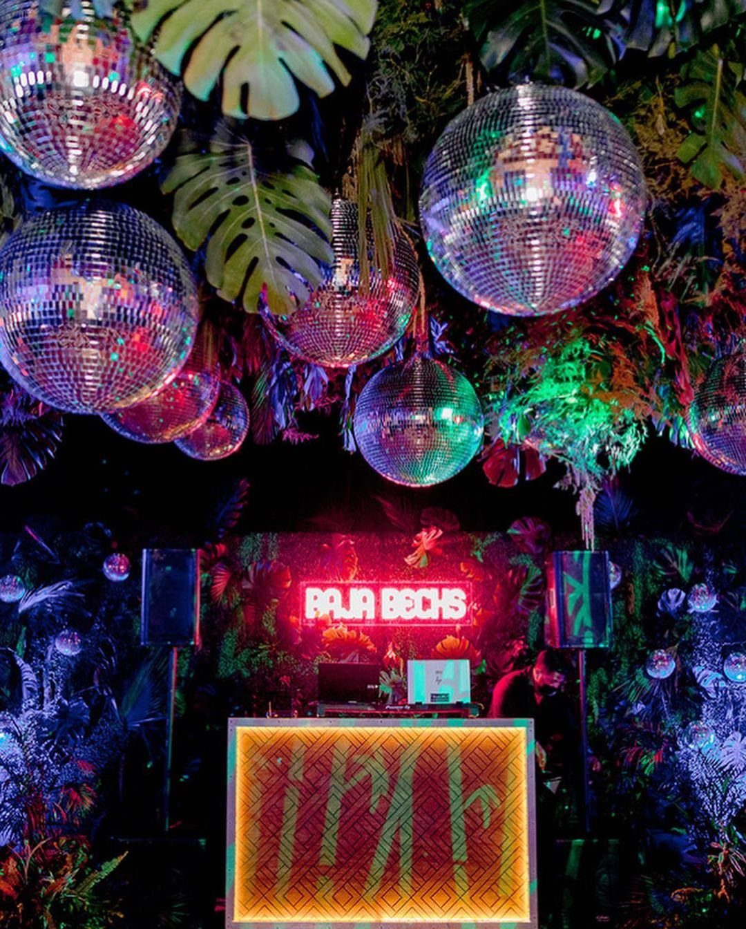 Disco balls hanging among tropical monstera leaves with a neon Baja Beachs sign.