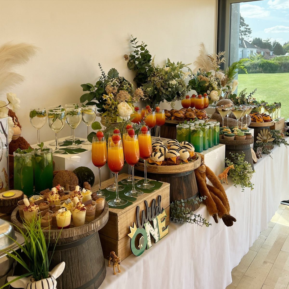 Safari party buffet table with orange and green drinks, desserts, and wooden barrels.