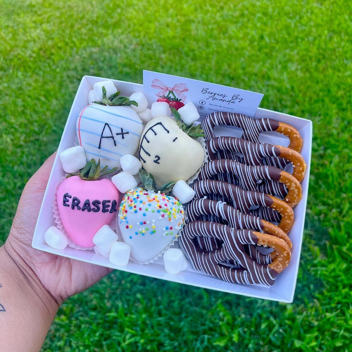 Decorated chocolate-dipped strawberries and pretzels in a white box held over green grass.