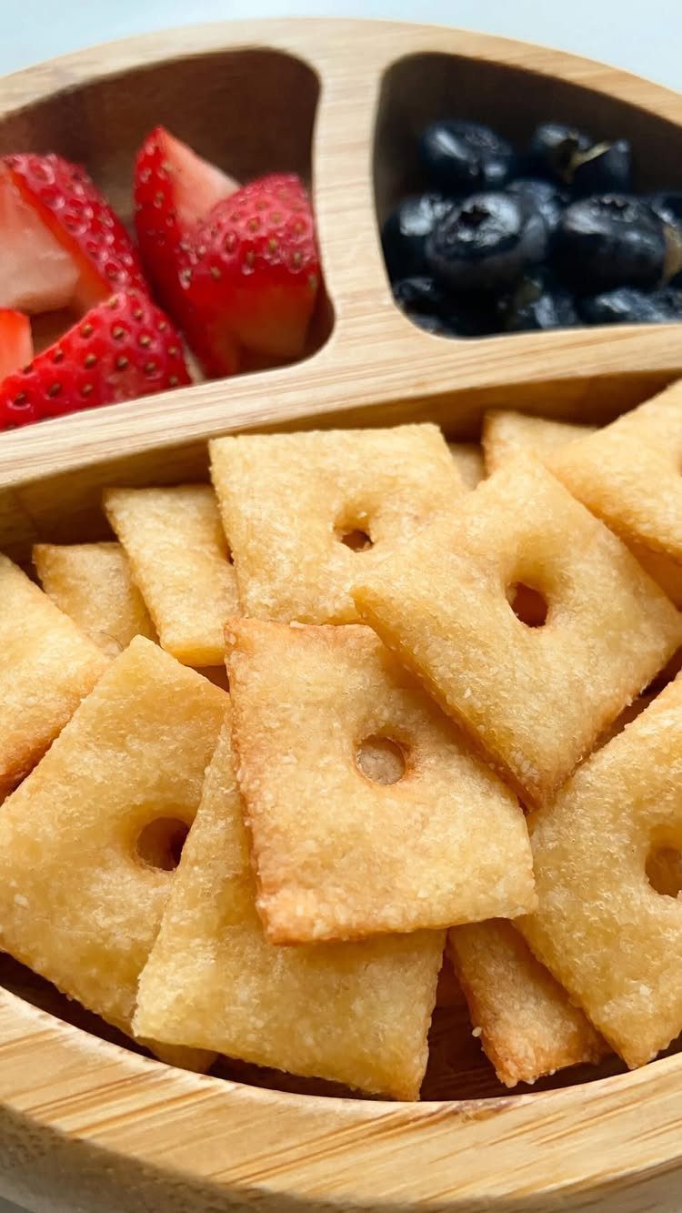 Square crackers, sliced strawberries, and blueberries in a wooden divided snack tray.