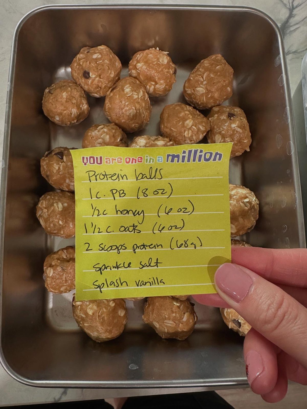 Hand holding a recipe note over a metal container of oat energy balls.