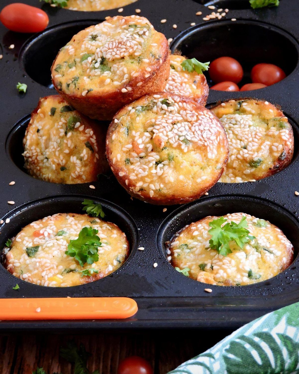 Savory mini muffins with sesame seeds in a black muffin tin with cherry tomatoes.