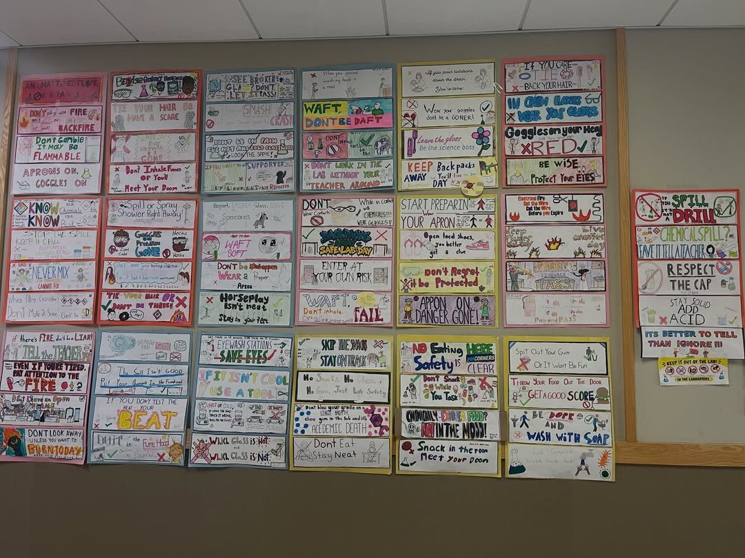 Student lab safety posters displayed in a grid on a classroom wall.
