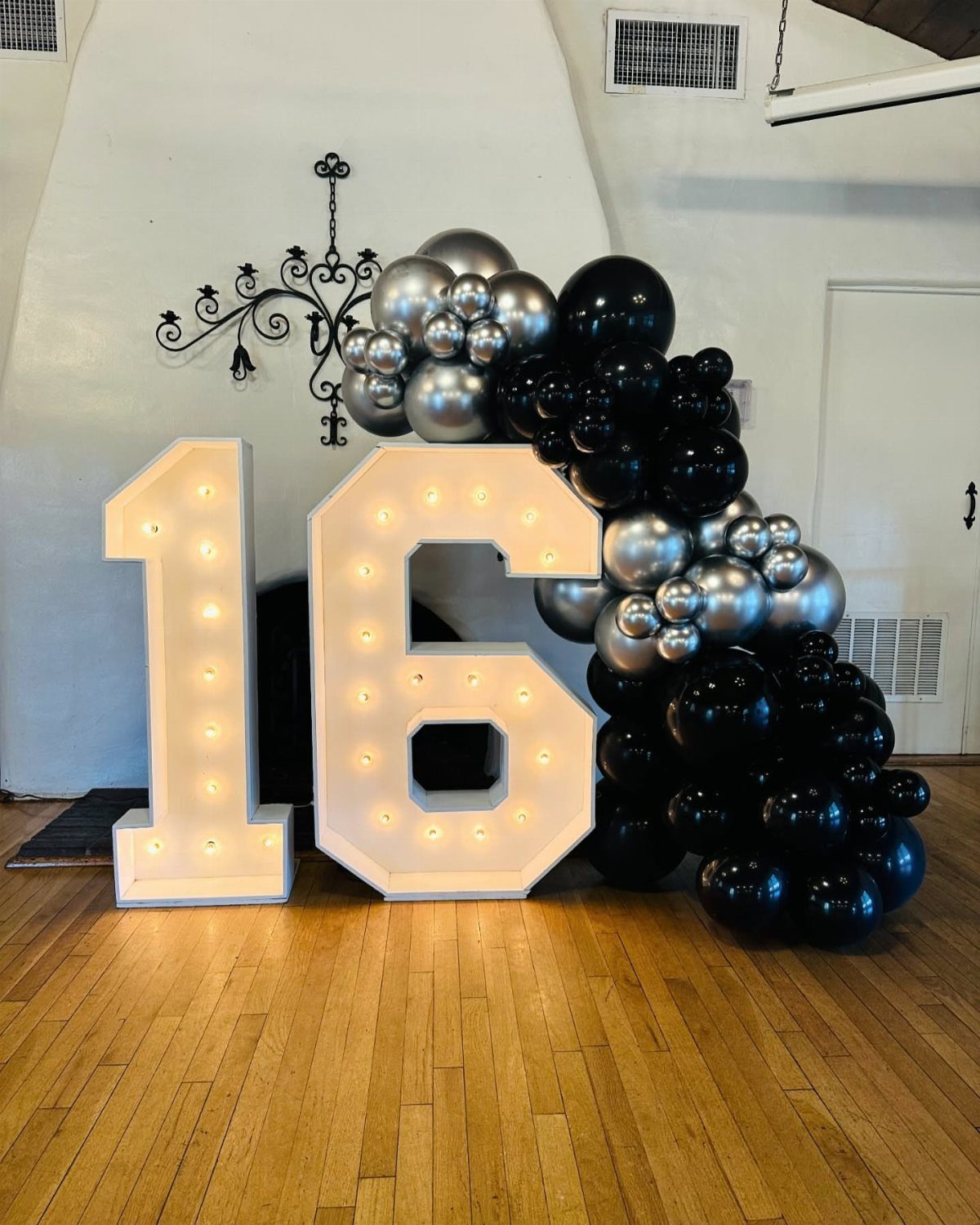 Large white illuminated marquee numbers 16 with black and silver balloon garland.