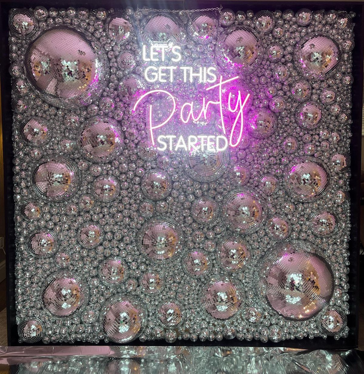 Silver sequin wall with disco balls and a pink neon party sign.