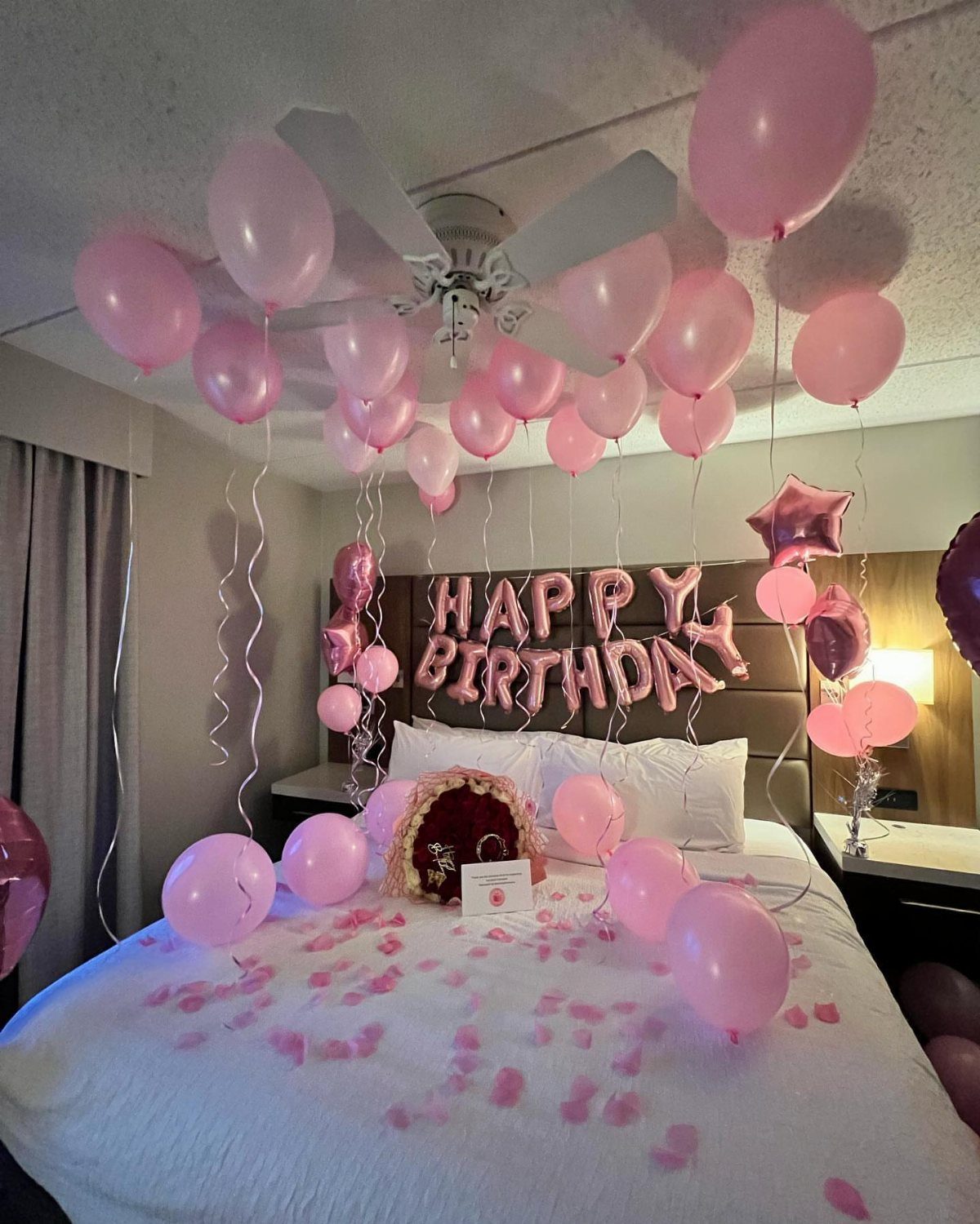 Pink birthday balloons and petals decorating a white bed in a bedroom.