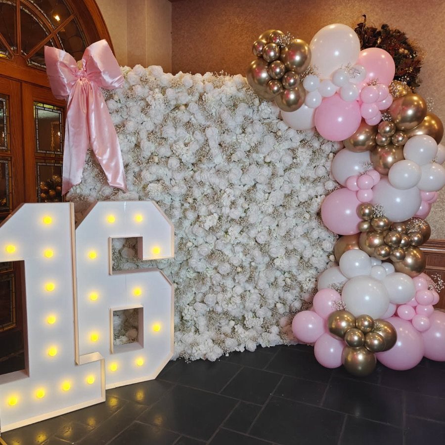 Illuminated marquee number 16 next to a white floral wall and pastel balloons.
