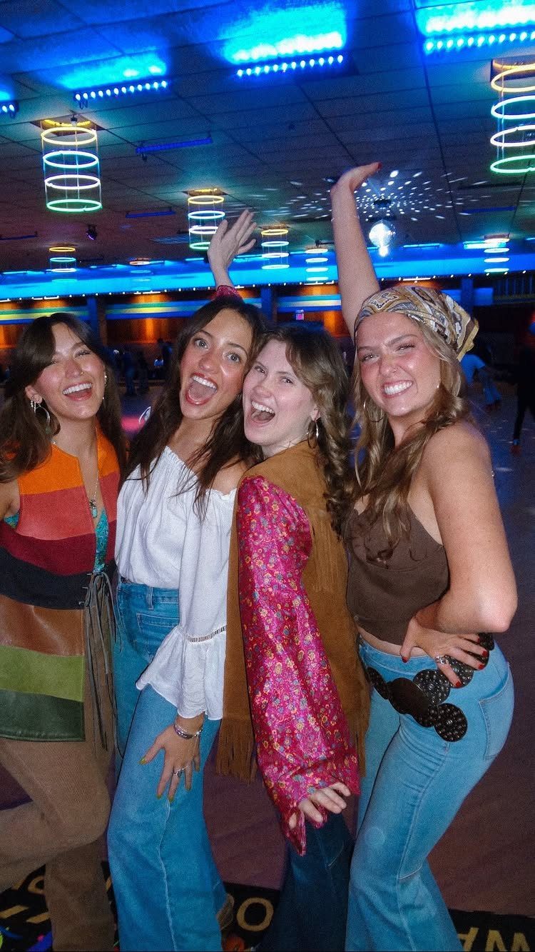 Four young women in 70s attire posing inside a neon-lit roller rink.