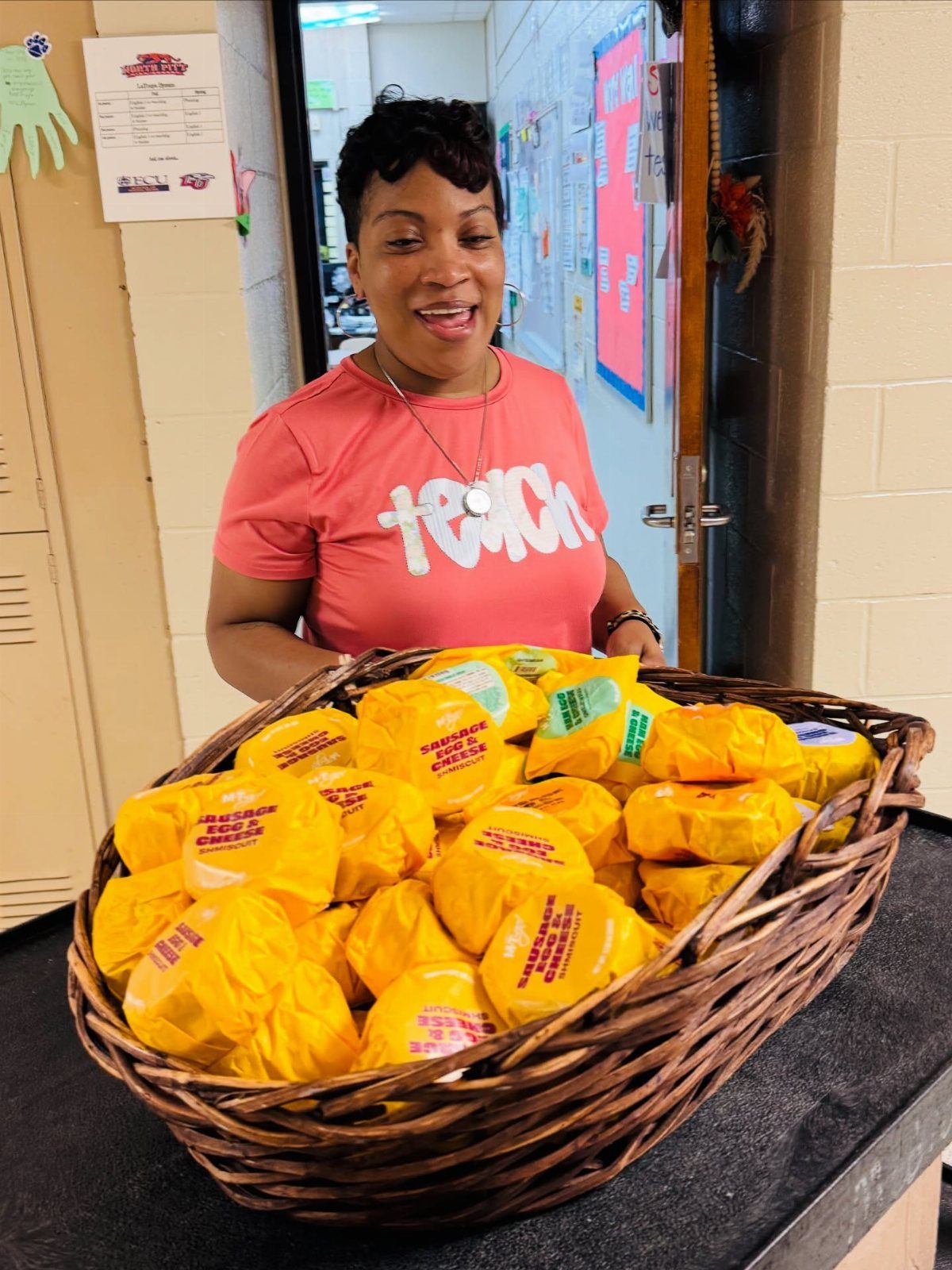 Woman in pink shirt standing behind a basket of wrapped breakfast sandwiches.