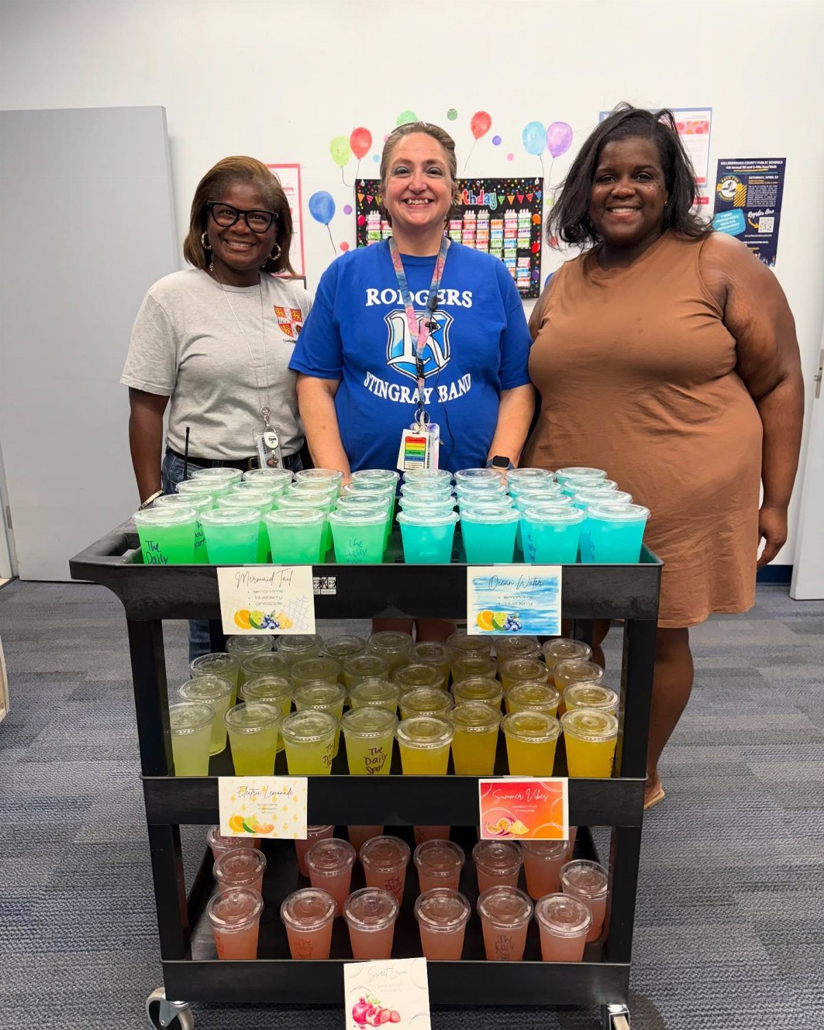 Three women standing behind a rolling cart filled with colorful smoothie cups.