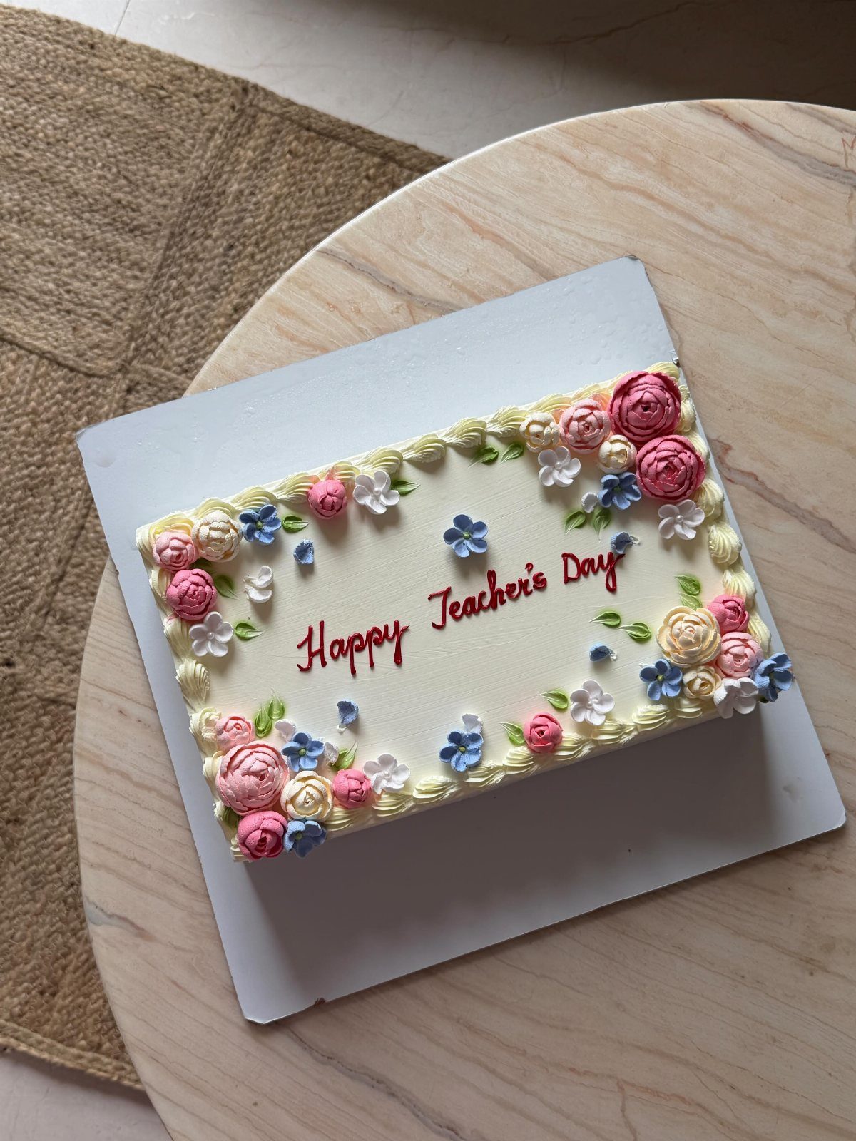 Rectangular white cake with colorful buttercream flowers and Happy Teacher's Day text.