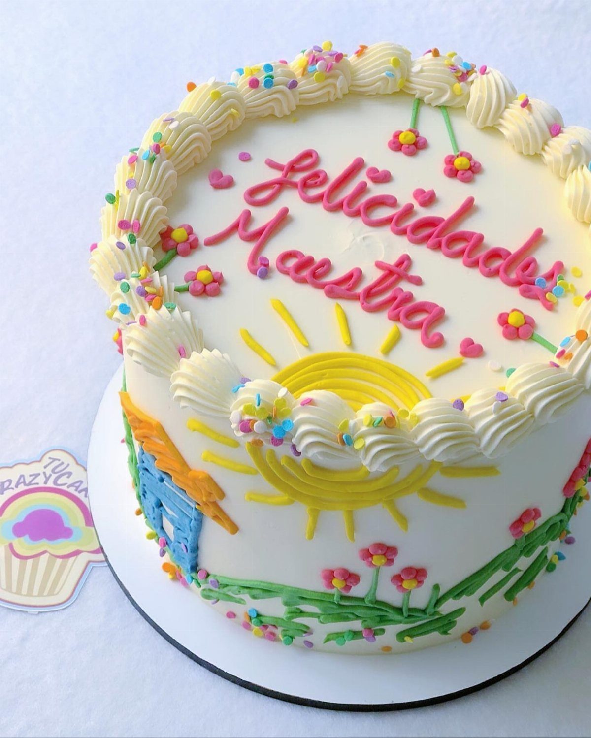 White frosted cake with a yellow sun and pink icing saying Felicidades Maestra.