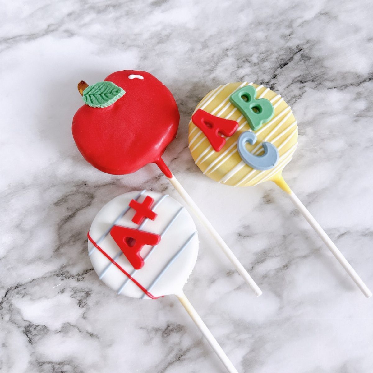 Three school-themed cake pops shaped like an apple, ABCs, and an A+ grade.