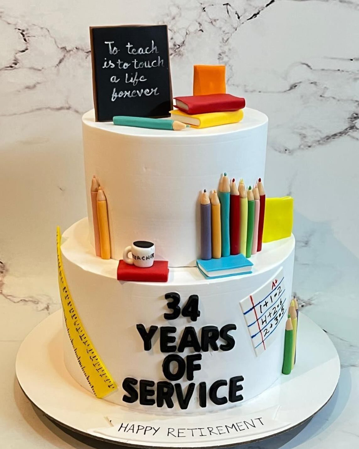 Two-tier white cake decorated with school supplies and text celebrating 34 years of service.